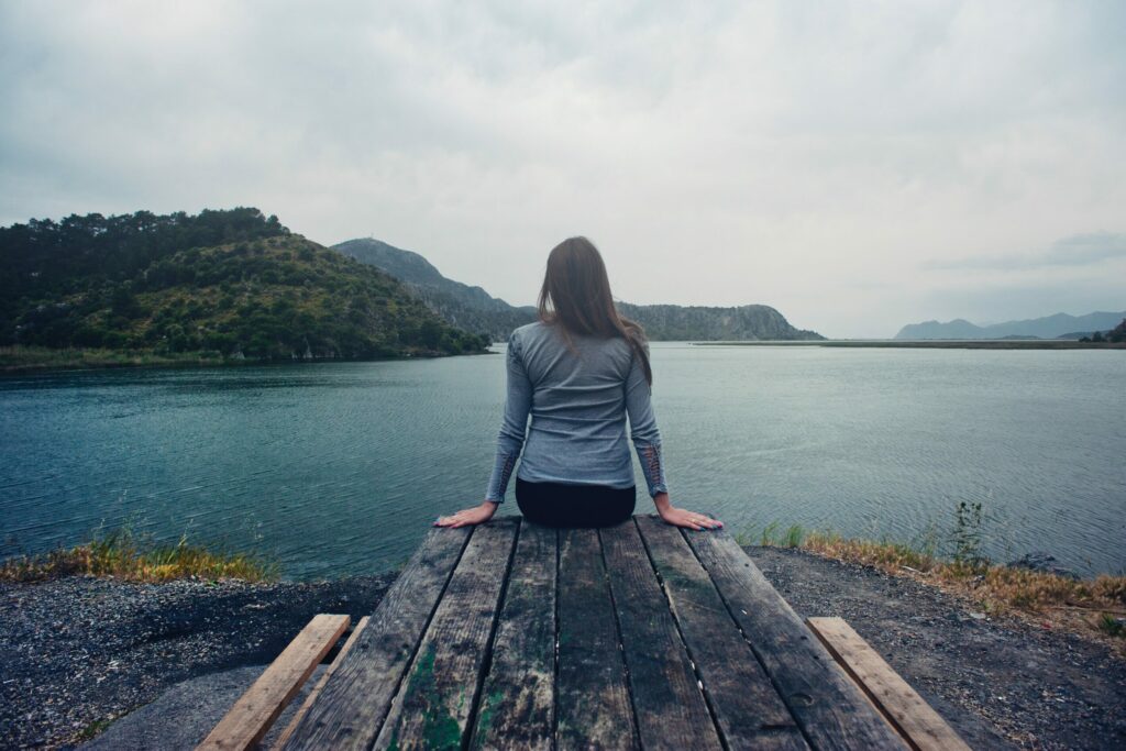 Femme assise au bout d'un quai qui admire le paysage composé de montagnes et d'étendue d'eau. Elle fait une rétrospective sur sa vie.