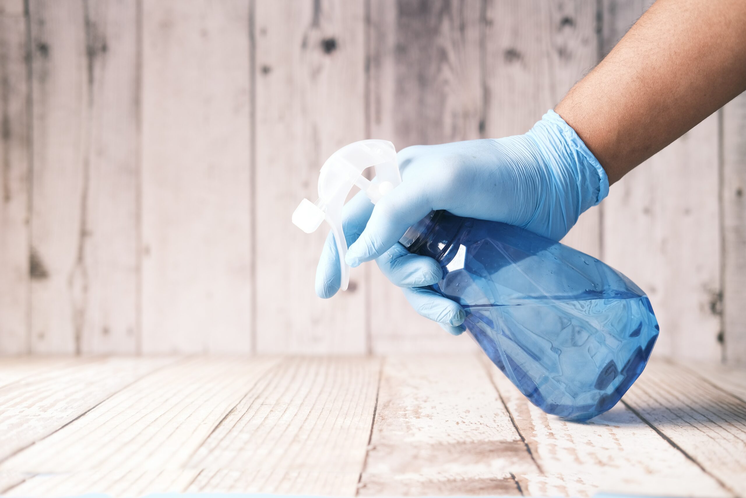 Image d'un produit nettoyant pour le bois avec une main recouverte par un gant en latex bleu sur fond de planches de bois.