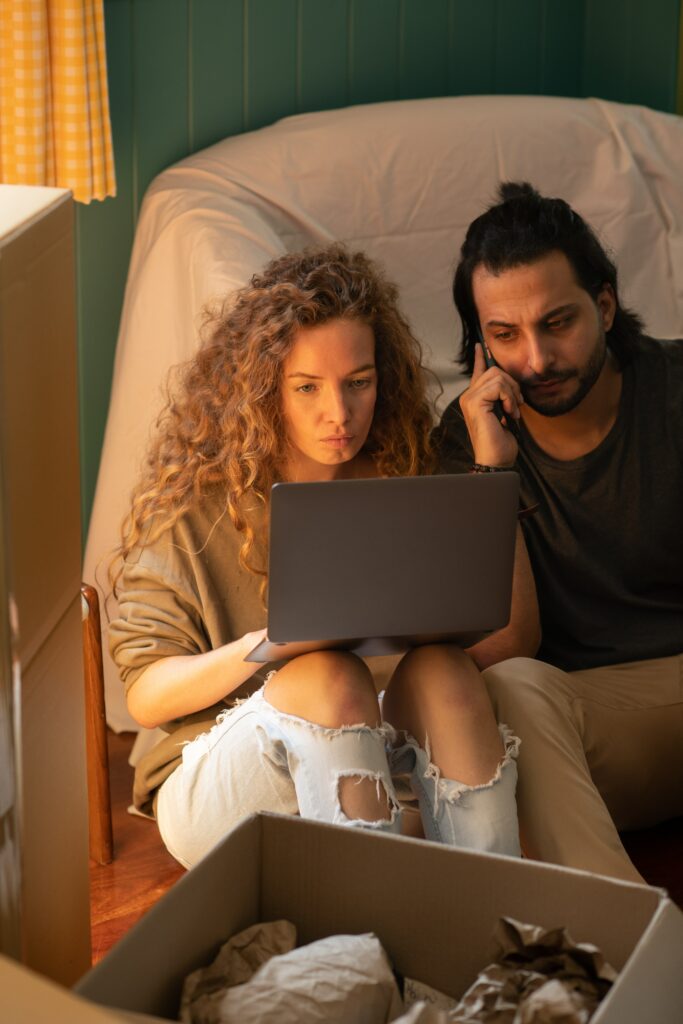 Un jeune couple constitué d'une femme et d'un homme qui sont assis par terre au milieu de leur déménagement effectue leurs changements d'adresse.