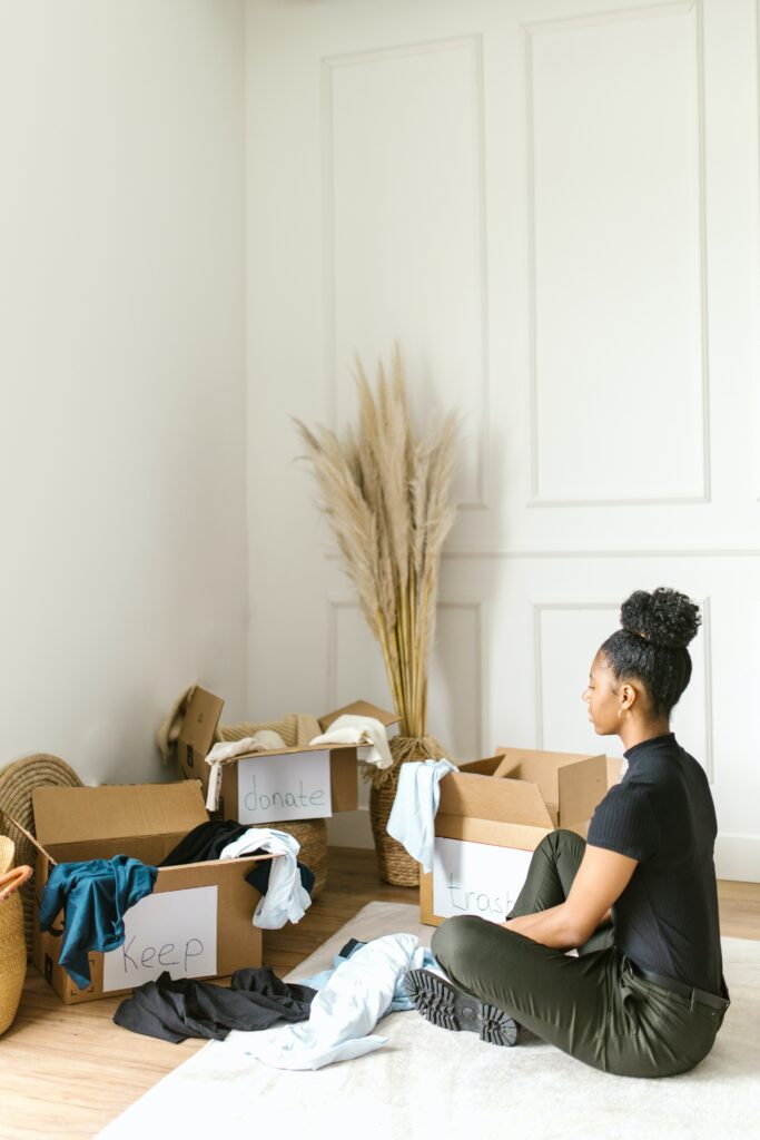 Jeune femme noire assise en indien par terre face à des boîtes en carton qui contiennent des biens triés par catégories. 