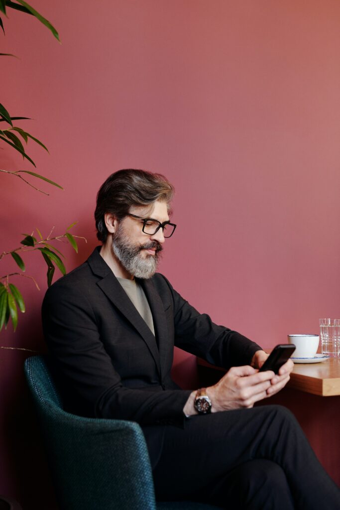 Homme d'affaire avec une barbe poivre et sel qui est assis dans un café et qui fait de la gestion sur son cellulaire.