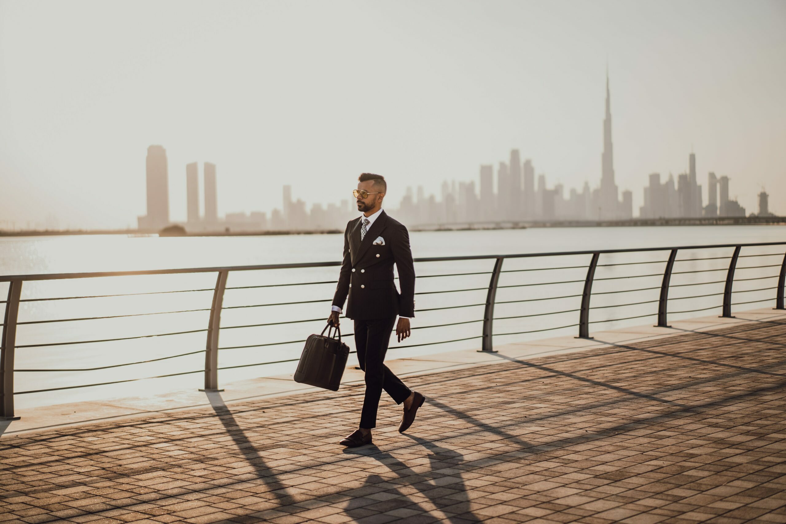 Homme d'affaire en complet et la mallette à la main, il est organisé dans sa vie. On le voit marchant sur un boardwalk avec un arrière plan d'étendue d'eau et d'une ville.