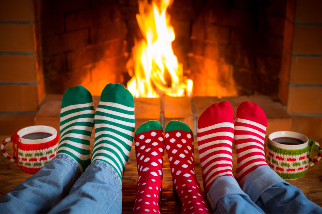 On voit les pieds d'adultes et d'enfants devant un feu de foyer et des tasses de chocolat chaud pendant le temps des fêtes. C'est le temps de prendre une pause en famille. 