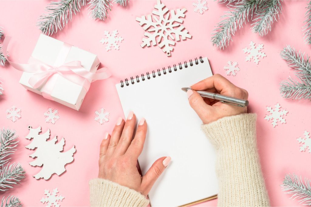 Planification du temps des fêtes. On voit la main d'une femme qui s'apprête à écrire une liste de choses à faire pour le temps des fêtes. La table sur laquelle elle écrit est parée d'une nappe rose et de décorations de Noël.