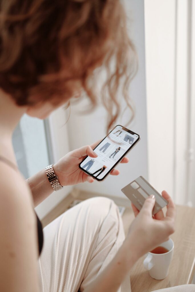 Jeune femme rousse aux cheveux courts qui fait des achats de vêtements sur son cellulaire et elle tient sa carte de crédit dans l'autre main.