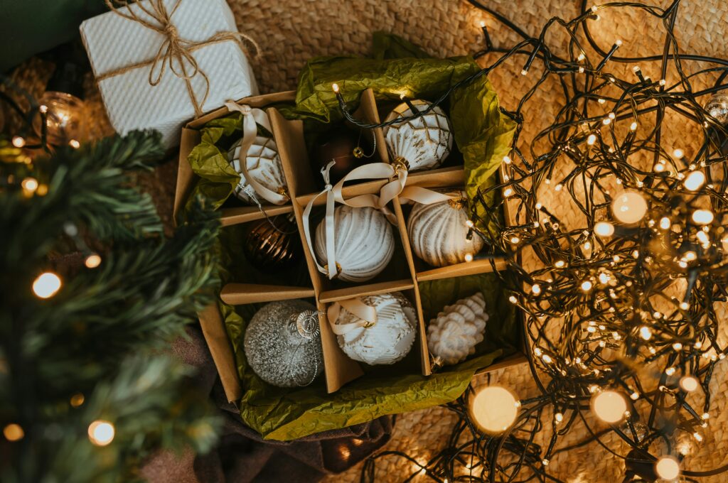 boîte compartimentée contenant de délicates décorations de Noël, soigneusement rangées avec du papier de soie vert pour les protéger. À côté, des guirlandes lumineuses dorées créent une ambiance chaleureuse et festive. En arrière-plan, un emballage cadeau blanc avec une ficelle en jute et des branches de sapin apportent une touche naturelle. Une scène évoquant la magie et l’organisation des fêtes.