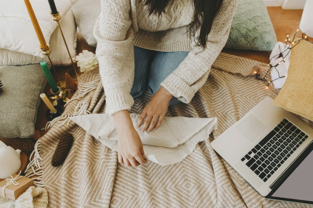 Une personne assise sur un tapis en laine texturée, portant un pull en tricot beige et un jean, prépare un emballage cadeau écologique en utilisant un tissu de type furoshiki. Autour d’elle, des coussins moelleux, des bougies décoratives, des guirlandes lumineuses et un ordinateur portable créent une ambiance chaleureuse et festive. Une scène qui évoque à la fois le confort et la créativité dans les préparatifs de Noël.