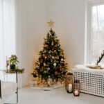 pièce lumineuse et minimaliste décorée pour Noël. Un sapin de Noël élégant, orné de boules dorées et blanches, est placé près d'une fenêtre avec vue sur un paysage hivernal. À droite, une banquette blanche recouverte d'une couverture en fausse fourrure accueille des lanternes et des bougies, ajoutant une ambiance chaleureuse. À gauche, un fauteuil en métal noir avec un plaid beige est accompagné d'une petite table décorée de verdure et de bougies. Une atmosphère apaisante et festive.