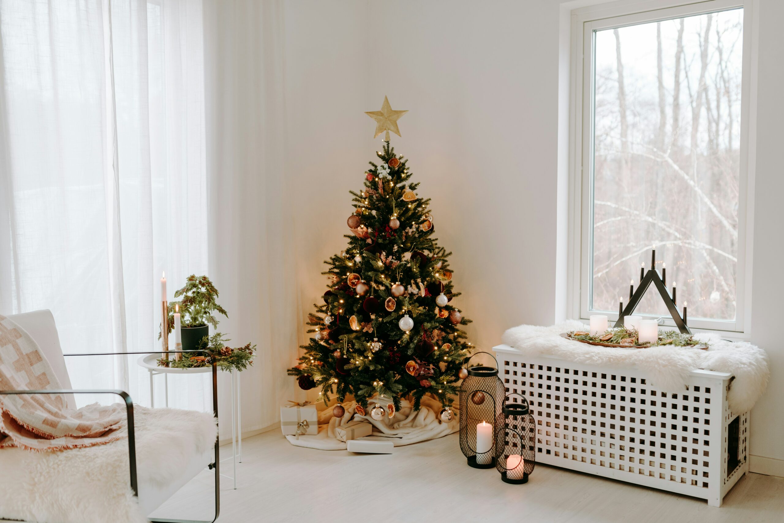 pièce lumineuse et minimaliste décorée pour Noël. Un sapin de Noël élégant, orné de boules dorées et blanches, est placé près d'une fenêtre avec vue sur un paysage hivernal. À droite, une banquette blanche recouverte d'une couverture en fausse fourrure accueille des lanternes et des bougies, ajoutant une ambiance chaleureuse. À gauche, un fauteuil en métal noir avec un plaid beige est accompagné d'une petite table décorée de verdure et de bougies. Une atmosphère apaisante et festive.