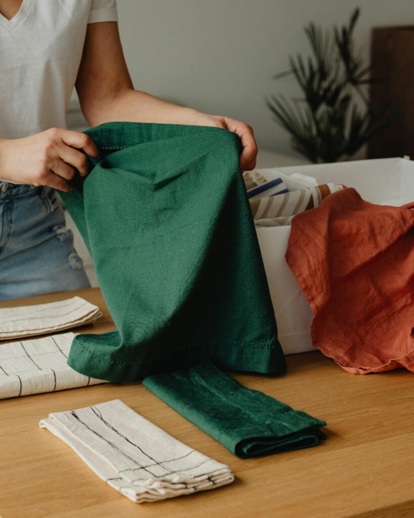 Une personne plie soigneusement une nappe verte sur une table en bois, entourée de serviettes de table en lin dans des tons neutres et d’une boîte contenant d’autres textiles, dont une nappe rouge. La scène évoque un moment d’organisation et de rangement, dans une ambiance calme et ordonnée. En arrière-plan, une plante apporte une touche de verdure à l’environnement.