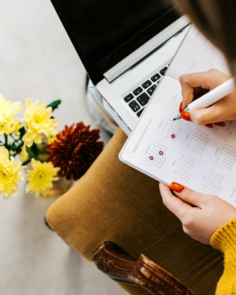 Une personne tient un agenda ouvert, entourée d'un ordinateur portable et de fleurs fraîches jaunes et rouges. Elle utilise un stylo pour entourer des dates importantes sur le calendrier de l'année, suggérant une planification ou des résolutions pour la nouvelle année. L’ambiance est lumineuse et organisée, évoquant la préparation et l’anticipation d’un futur bien structuré.