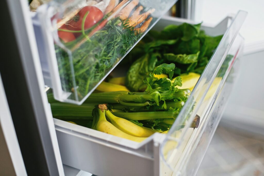 Des tiroirs de réfrigérateur bien organisés remplis de légumes et de fruits frais. Le premier tiroir contient des tomates, des carottes et des herbes comme le persil, tandis que le deuxième tiroir regroupe des bananes, du céleri et de la laitue. Une disposition fonctionnelle qui facilite l'accès aux aliments et prolonge leur fraîcheur.