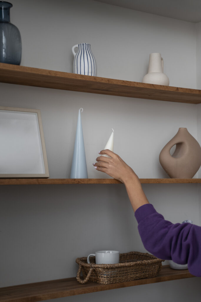 Une main ajuste une bougie décorative sur une étagère en bois, illustrant un détail soigné dans un projet de valorisation immobilière.