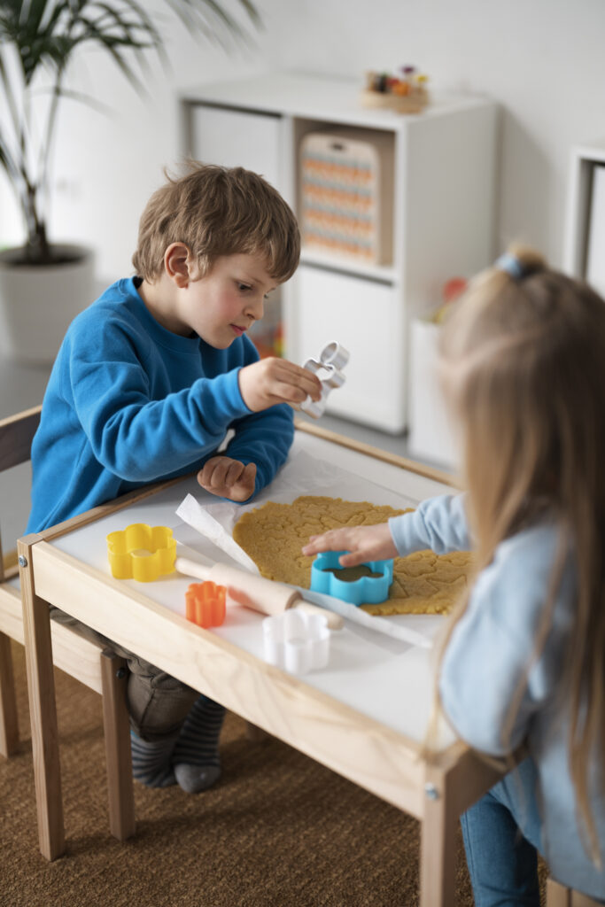 Deux enfants découpent des formes dans de la pâte à modeler sur une petite table, illustrant un moment de jeu créatif rendu possible grâce à une organisation de la salle de jeux bien pensée et adaptée à leur autonomie.