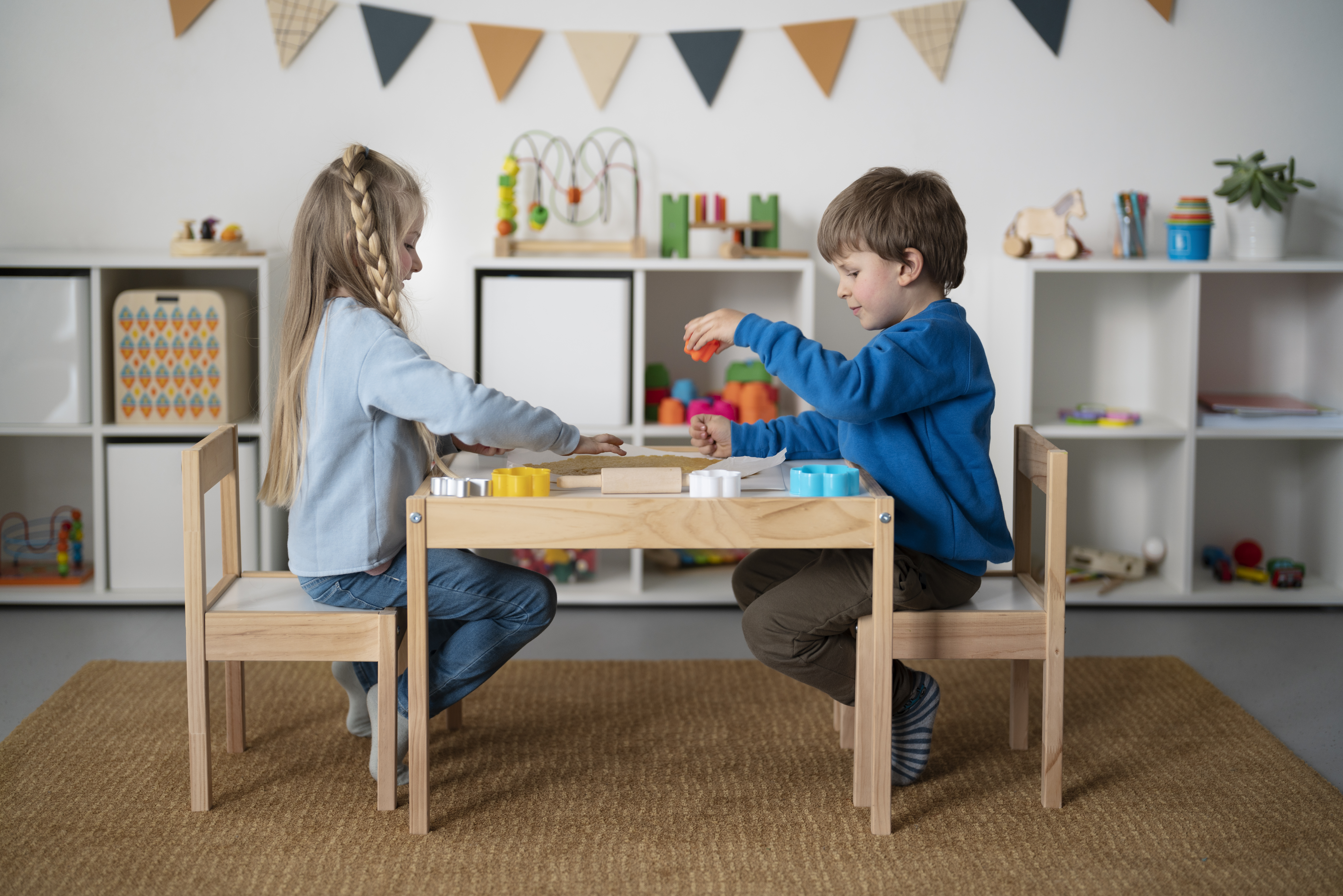 Deux jeunes enfants jouent ensemble à une table basse dans une pièce lumineuse, illustrant une organisation de la salle de jeux bien pensée, avec des bacs de rangement accessibles et des jouets soigneusement disposés en arrière-plan.