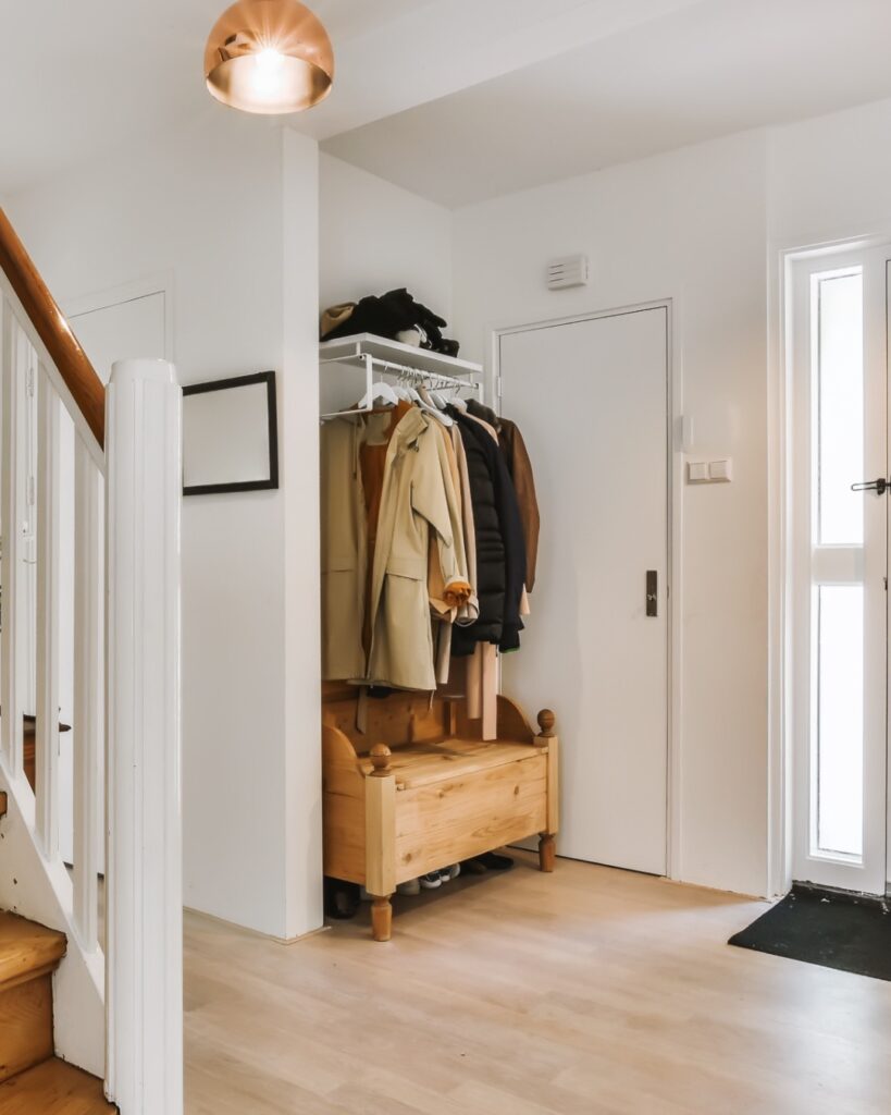 Entrée de maison bien rangée avec banc de rangement en bois, manteaux suspendus et tapis à la porte, baignée d’une lumière naturelle.
