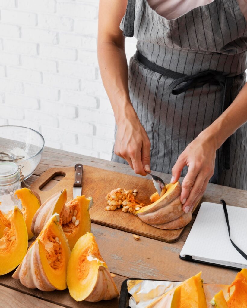 Personne en tablier en train de couper une courge sur une planche de bois dans une cuisine lumineuse, avec plusieurs morceaux de courge autour.