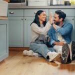 Couple souriant assis sur le plancher de la cuisine, se tapant dans la main dans un moment complice.