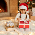 Petite fille assise devant un foyer décoré pour Noël, ouvrant un cadeau, une scène qui illustre l’importance du désencombrement avant l’arrivée des Fêtes.
