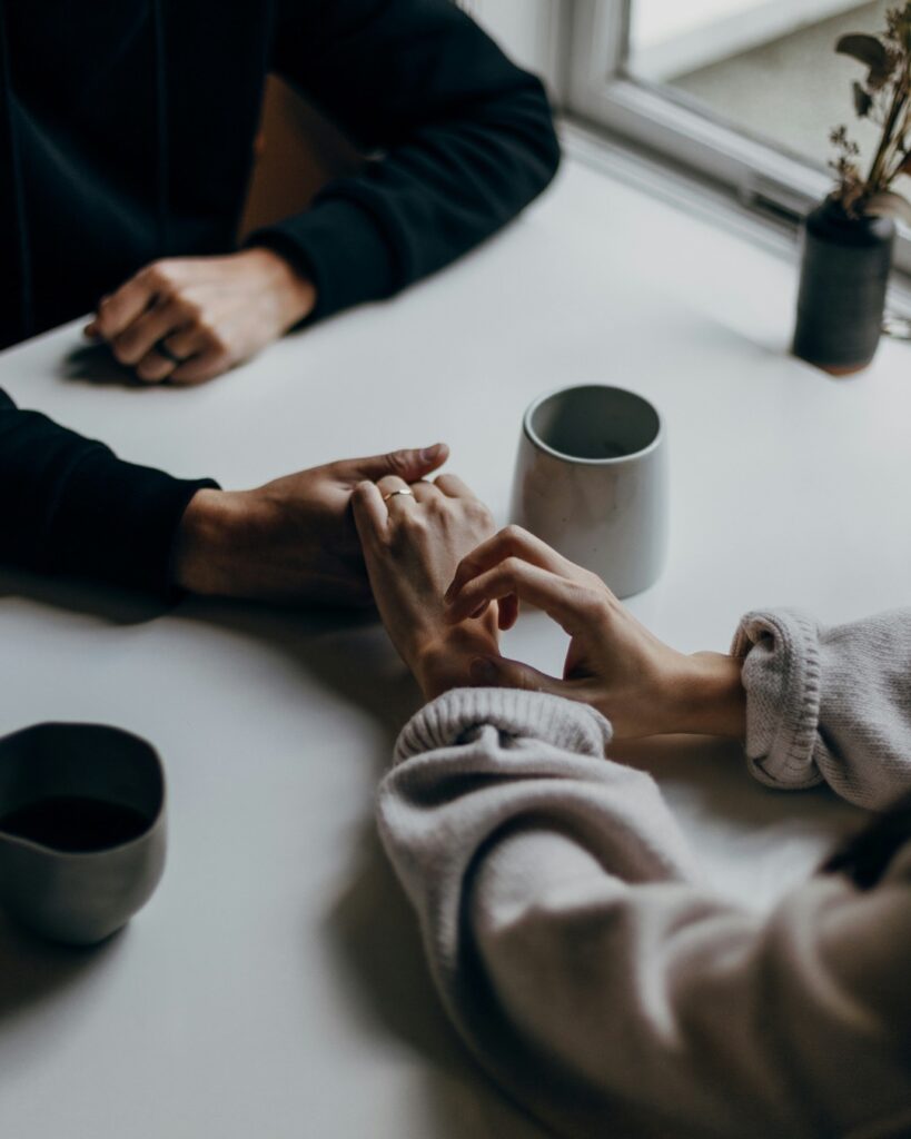Deux personnes assises à une table se tenant les mains, illustrant le soutien, l'accompagnement et l'entraide lors d'une période de transition personnelle ou familiale.